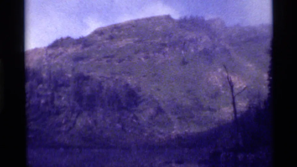 The Holland Lake mountain slope remains in view, with dark forest, rocky openings, and a purple-tinted 8mm home-movie look.