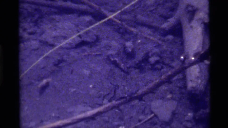 1972 8mm underwater view at Holland Lake, Montana, showing silty bottom texture, crossed twigs, and a pale stick along the right edge.