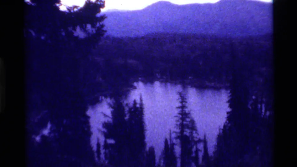 1972 Holland Lake, Montana view of water framed by evergreen trees, with wooded hills and hazy mountains in blue-purple aged film color.