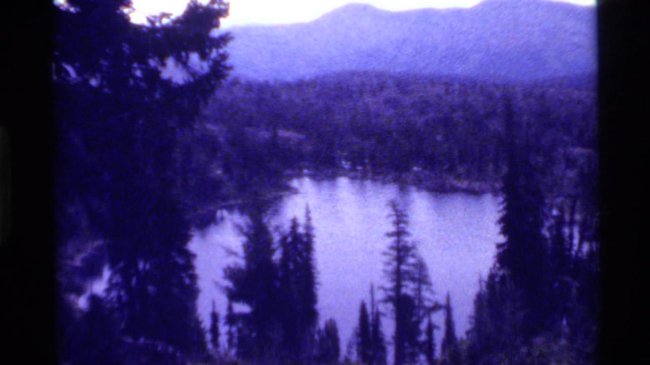 Calm lake below dark pine trees with forest and mountain ridges in the distance.
