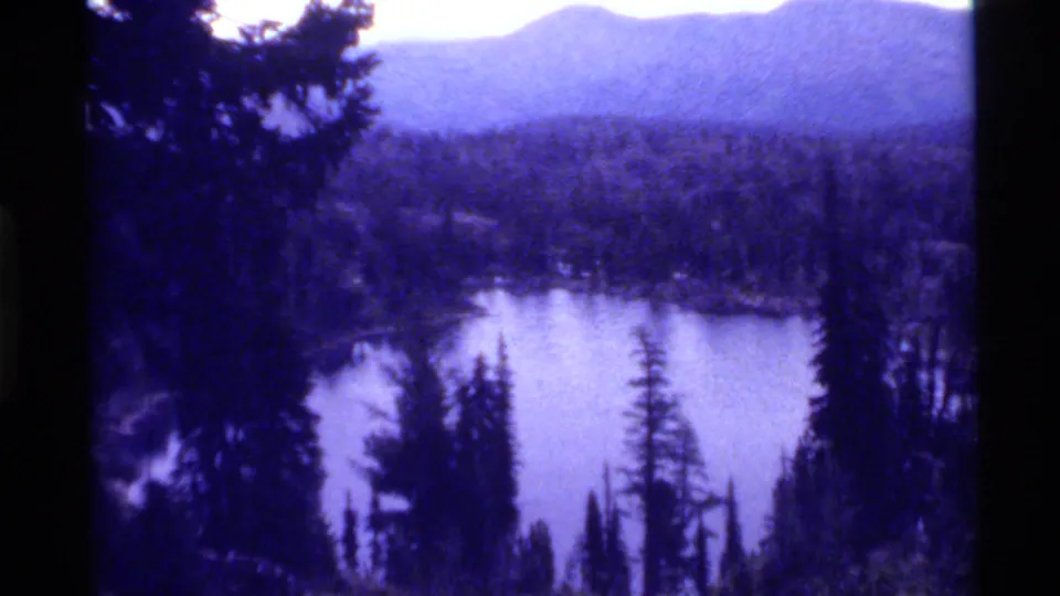 1972 8mm view of Holland Lake, Montana, with dark pine trees framing calm water, forest, and distant mountain ridges.