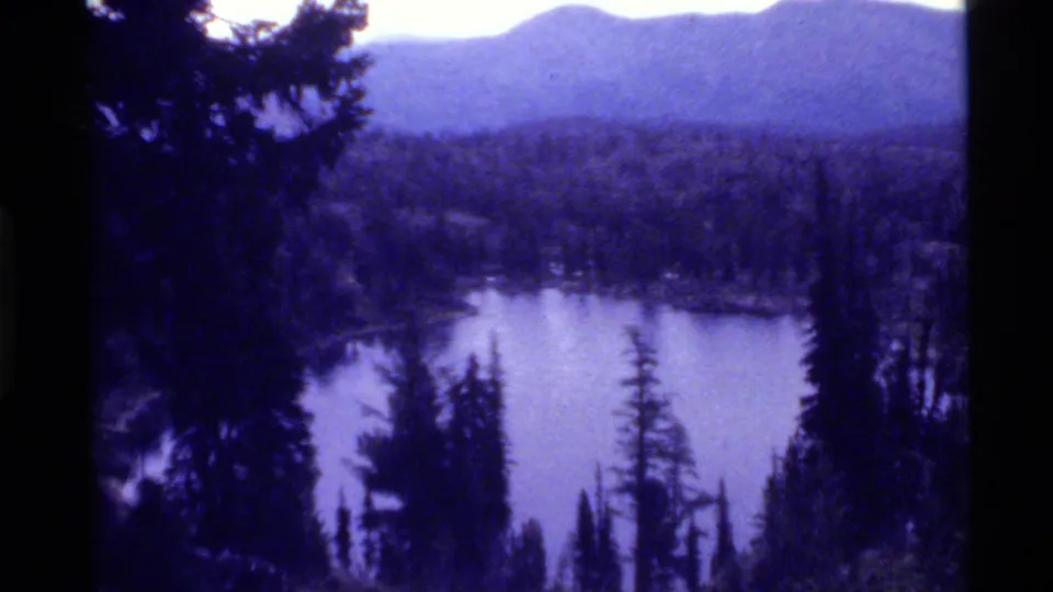 1972 8mm Holland Lake scene with still water, surrounding forest, dark foreground trees, and soft mountain silhouettes.