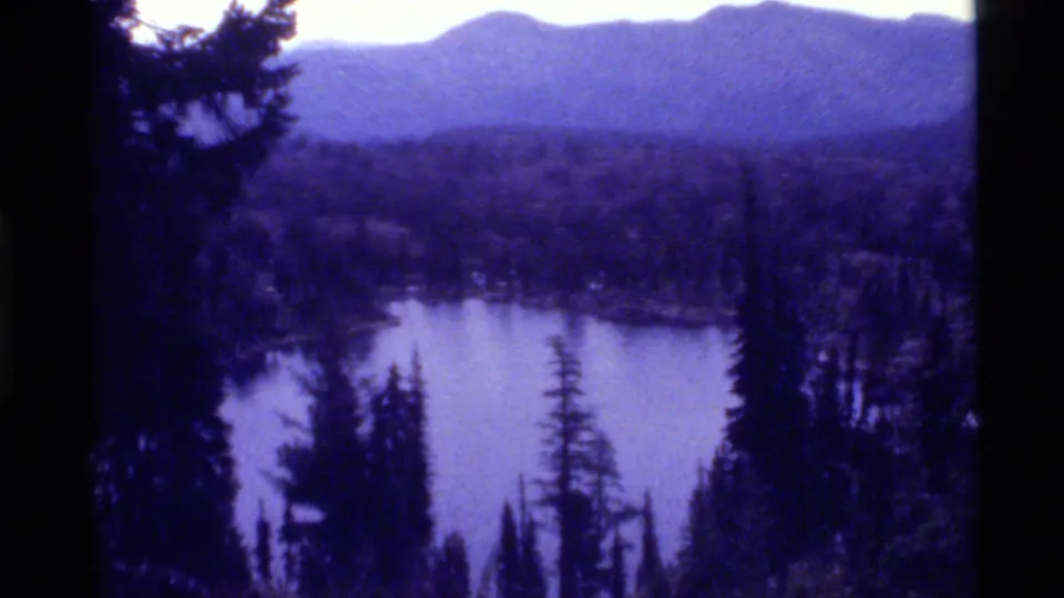 1972 home-movie frame from Holland Lake, Montana, showing calm water, pine silhouettes, forest, and distant mountains.