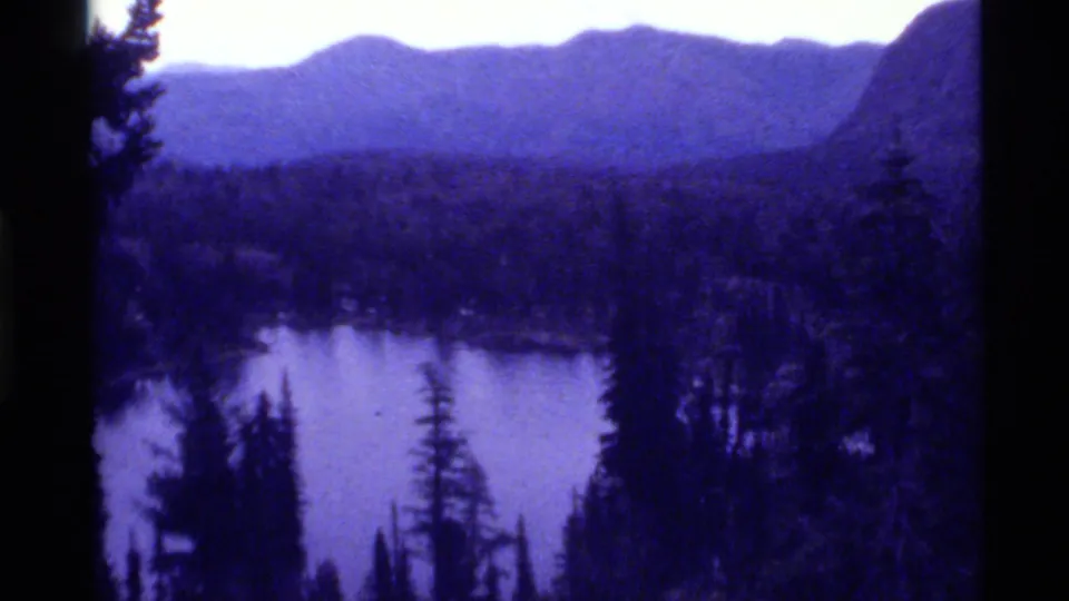 1972 8mm view of Holland Lake, Montana, with evergreen forest, distant mountains, pale sky, and a purple-blue film cast.