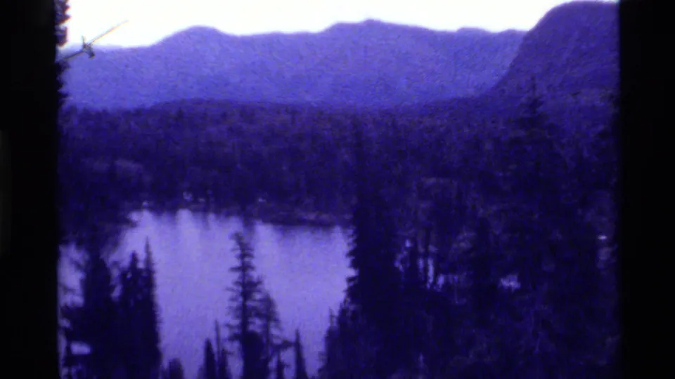 Wide 1972 8mm landscape of Holland Lake, Montana, framed by forest and distant mountain ridges with visible grain and edge shadow.