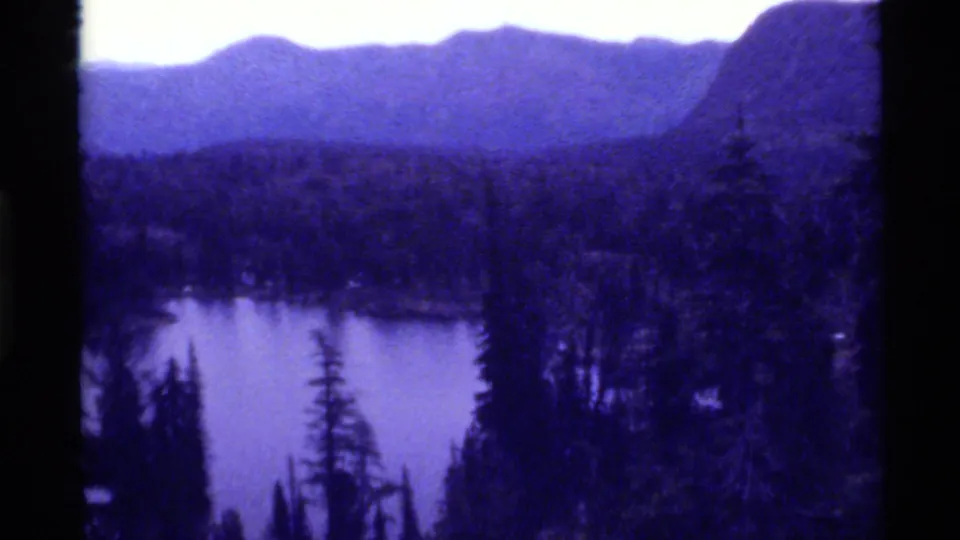 1972 8mm frame of Holland Lake with foreground evergreens, forested shoreline, and distant Montana mountains softened by film grain.