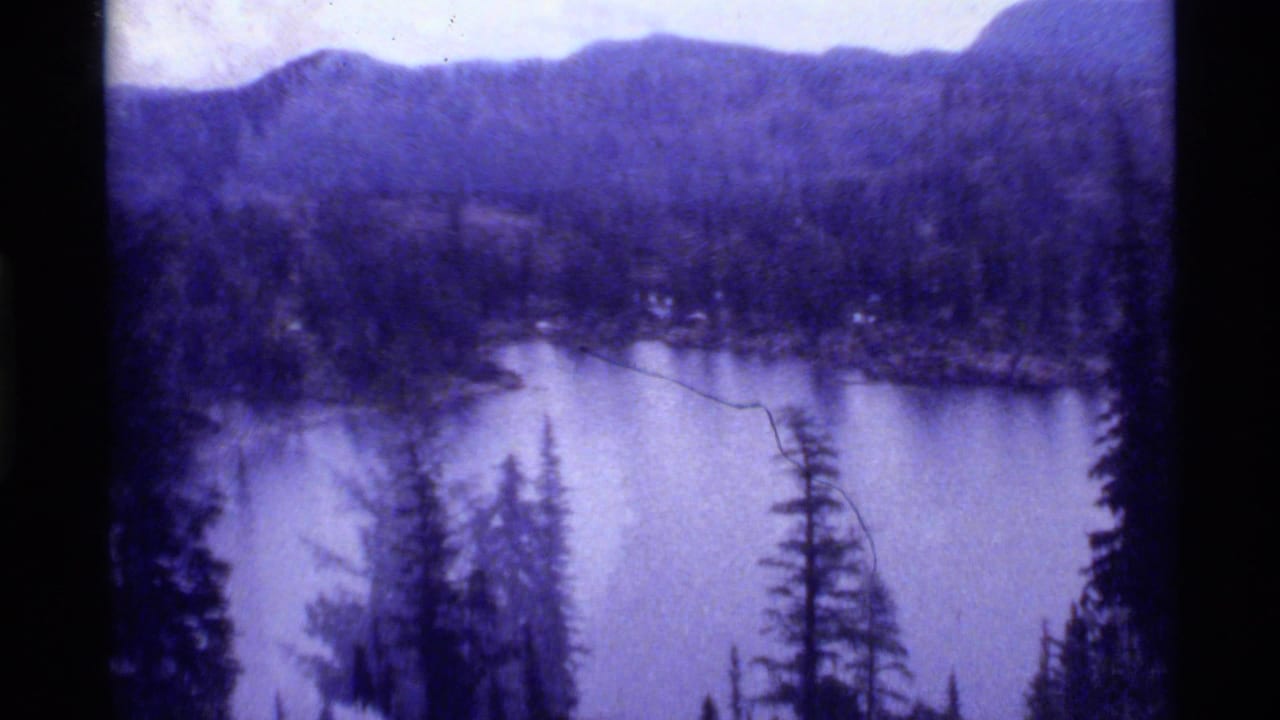 1972 8mm view of Holland Lake, Montana, framed by evergreen trees with wooded hills and distant mountains under cloud cover.