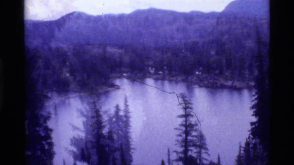 1972 8mm view of Holland Lake, Montana, framed by evergreen trees with wooded hills and distant mountains under cloud cover.