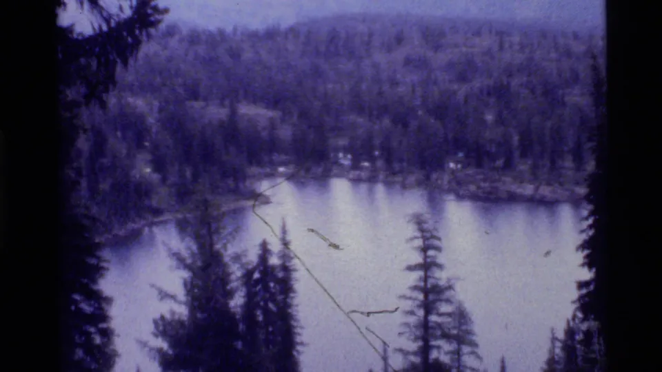 1972 8mm landscape view across Holland Lake toward evergreen shoreline, wooded hills and mountains, with visible aged-film scratches.