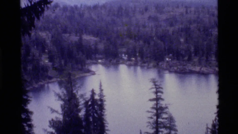 1972 8mm view of Holland Lake with dark foreground evergreens, pale water, wooded shore and softened mountain forms in the background.