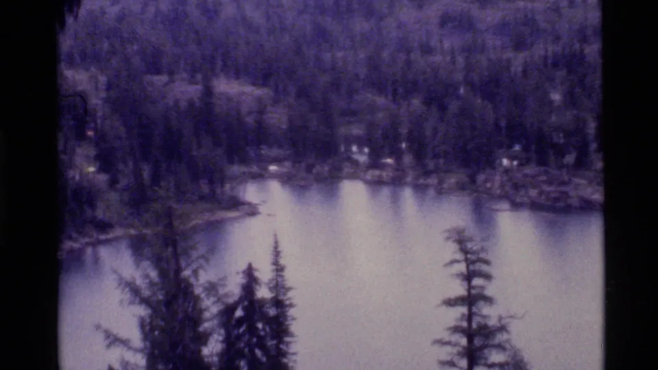 1972 8mm Holland Lake view from among evergreens, showing calm water, a wooded shoreline and distant hills in muted cloudy light.