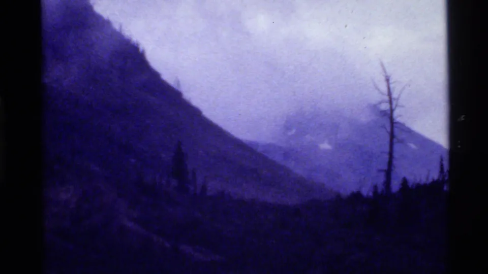 1972 Holland Lake, Montana frame with a steep shadowed slope, misty distant ridge, dark forest line, and bare tree at right.