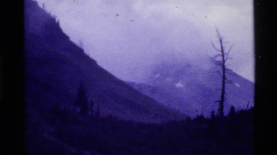1972 Holland Lake, Montana home-movie view of a foggy mountain ridge, dark treeline, steep left slope, and isolated bare tree.