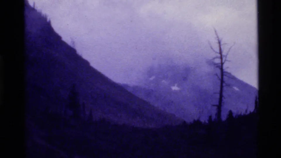 1972 Holland Lake, Montana frame showing a purple-blue mountain landscape, low cloud, forested foreground, and a bare tree near the right side.