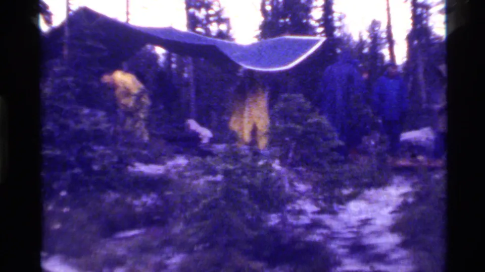 1972 Holland Lake campsite with a sloped tarp, yellow tent, pine trees, rocky ground, and indistinct people near camp.