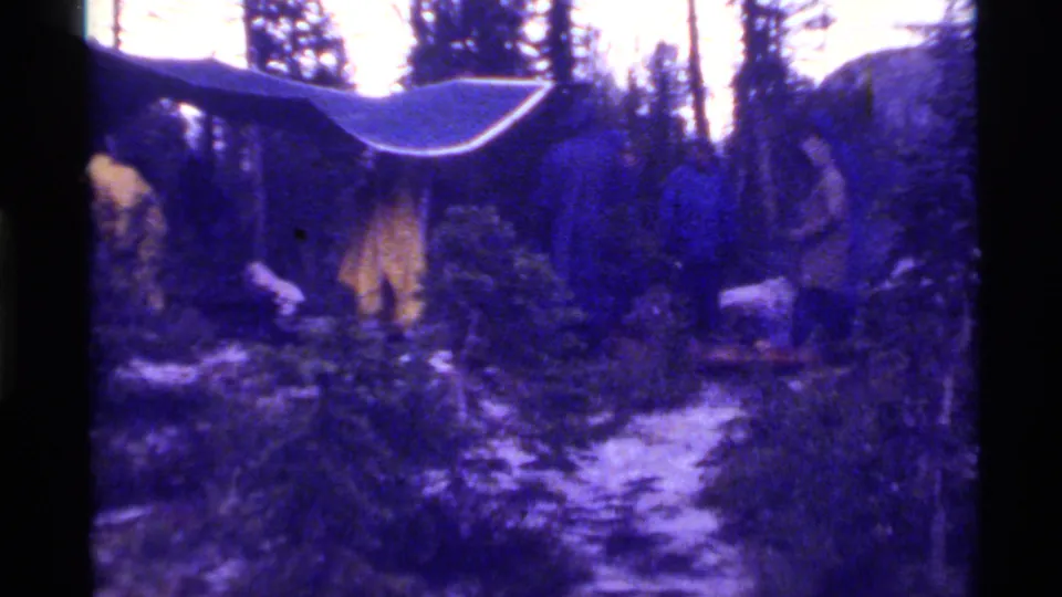 Home-movie frame of people gathered near a tarp shelter and yellow tent at Holland Lake, Montana, with trees and rocky terrain around them.