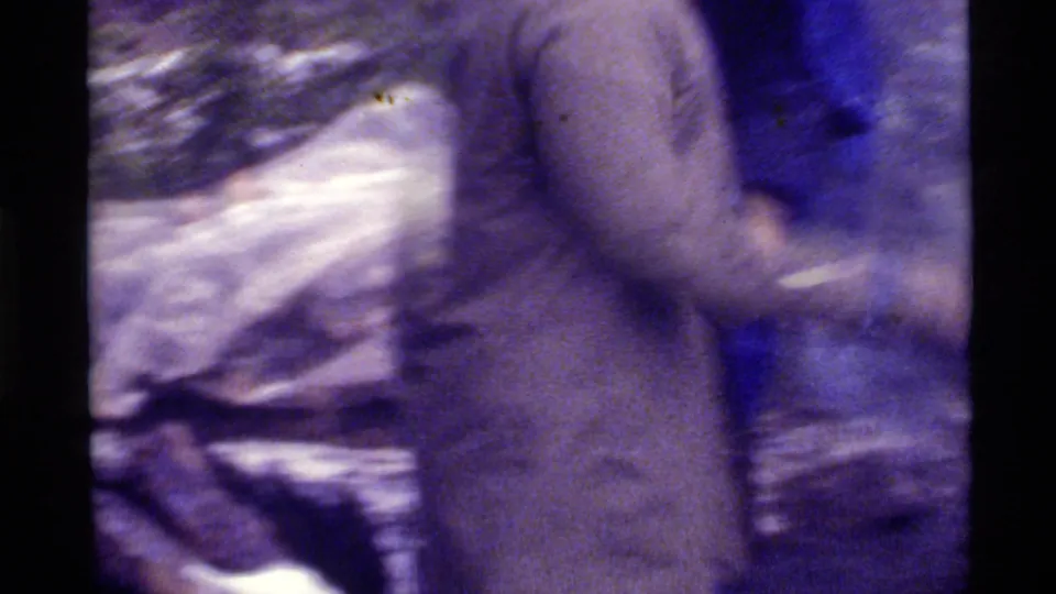 1972 Holland Lake, Montana frame showing a bundled person in a light coat moving through a snowy forest scene on aged 8mm film.
