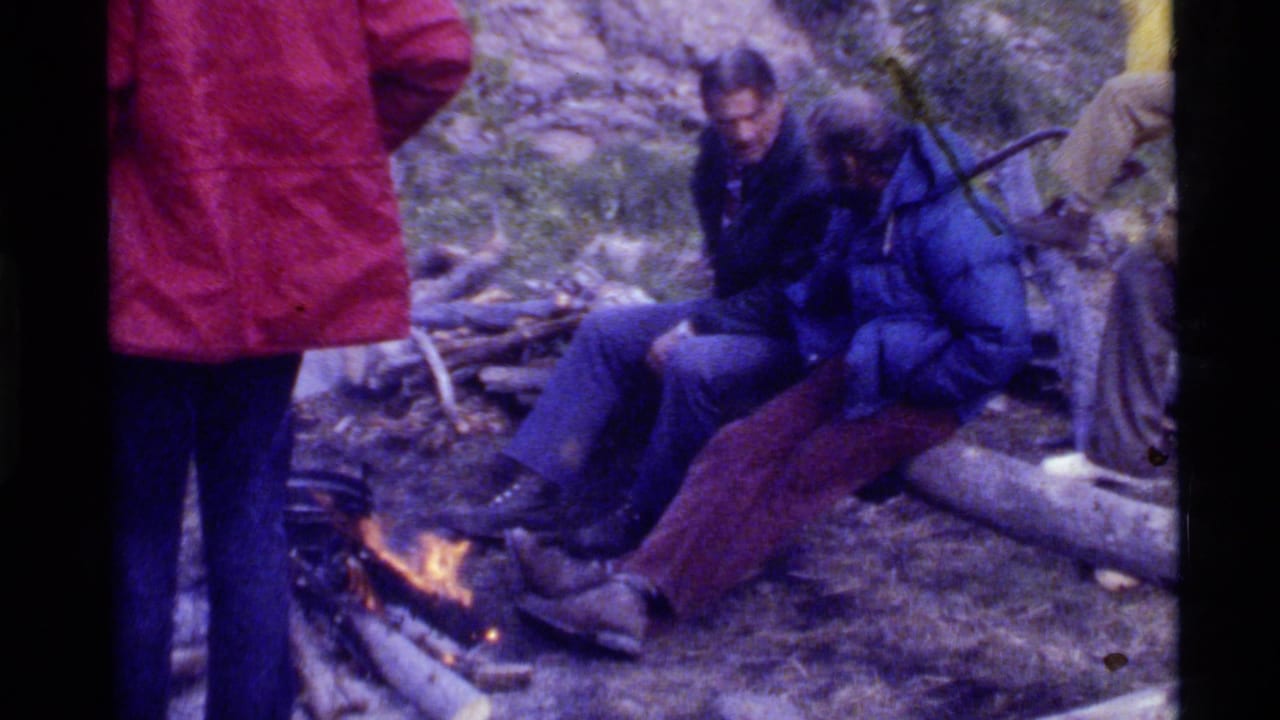At Holland Lake, Montana in 1972, campers gather by a small fire, with two people seated on logs and others standing nearby.