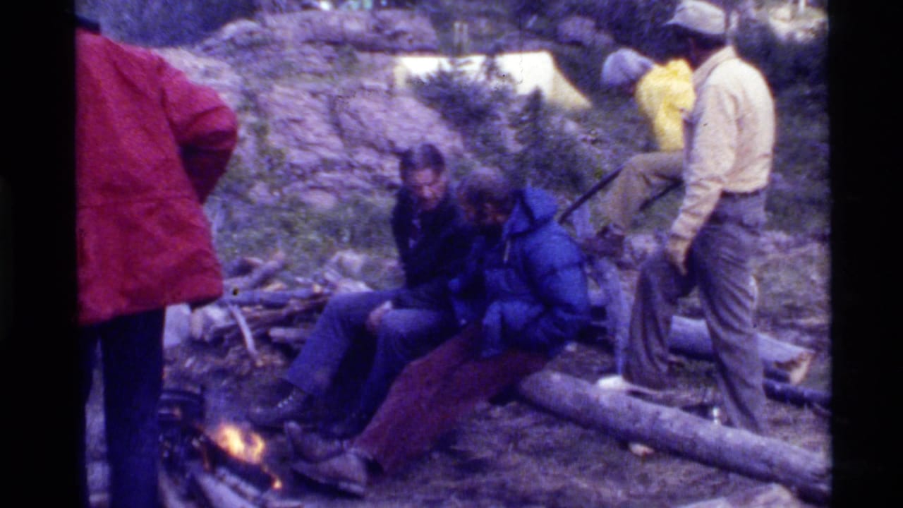 Several campers are gathered near a fire, with tents and brush visible behind them.