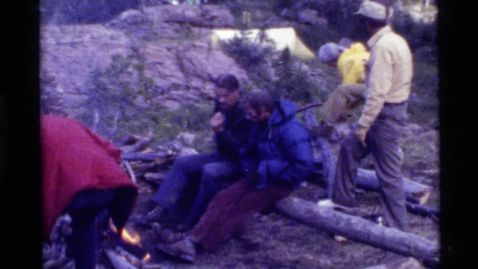 At the Holland Lake campsite, friends gather around the campfire as a seated man leans forward and others stand nearby.