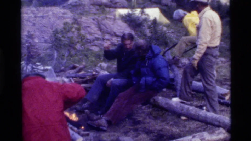 In the final frame, the Holland Lake camping group stays near the fire while one seated camper gestures beside the log seating.