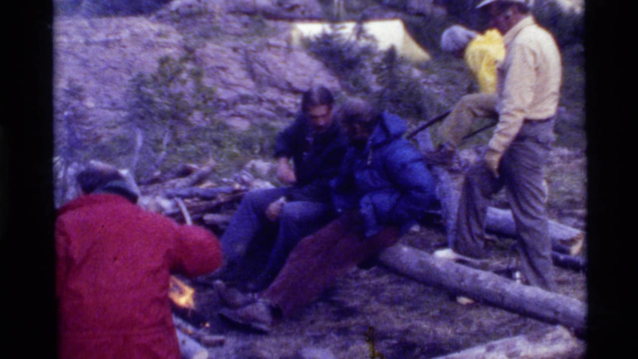 1972 Holland Lake home-movie frame showing men in red, blue, and light jackets gathered around a small campfire among logs and rocks.
