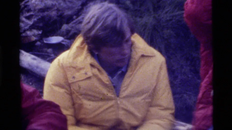 1972 Holland Lake frame with a man in a yellow insulated jacket seated near logs and brush, with other campers partly visible.