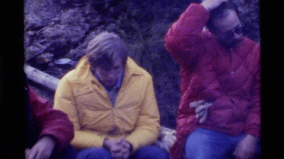 1972 Holland Lake home-movie frame showing men seated on logs, including one in a yellow jacket and one in red adjusting his headwear.