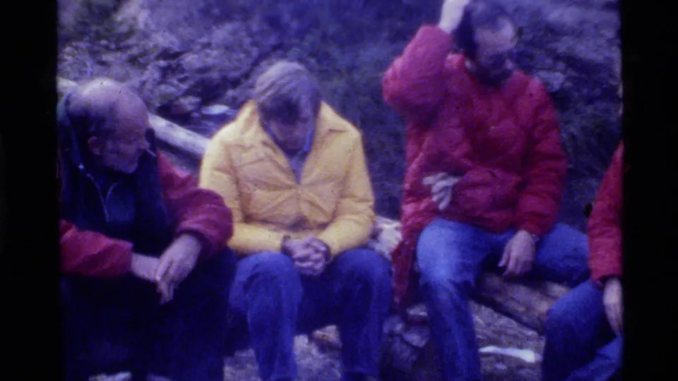 1972 Holland Lake frame with several adult men seated on logs in colorful jackets during a quiet wooded campsite gathering.