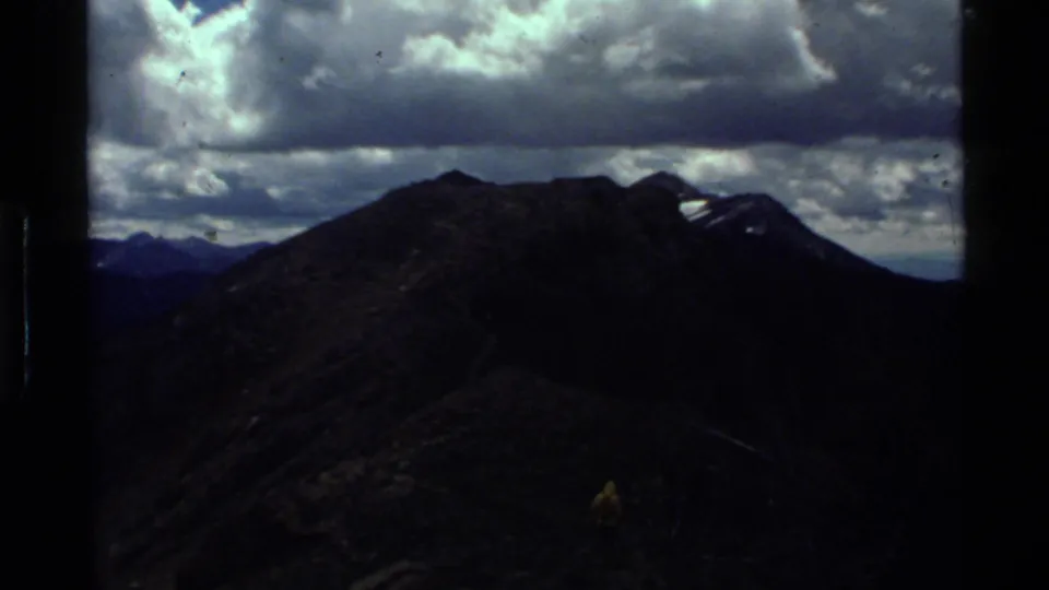 1972 Holland Lake, Montana home-movie frame showing a shadowed mountain ridge, cloudy sky, and a small distant hiker on the trail.