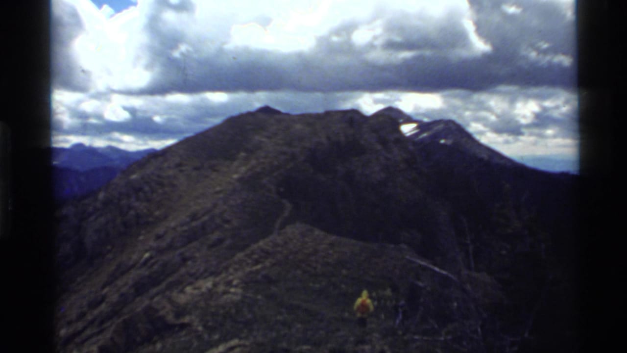 Mountain ridge and winding trail with a small hiker in a yellow jacket.
