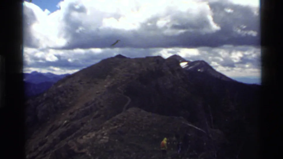 1972 Holland Lake, Montana home-movie view of a rugged mountain slope with a winding path and a distant hiker below bright clouds.