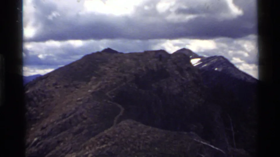 1972 Holland Lake, Montana frame showing a broad ridge panorama, winding mountain trail, distant peaks, and heavy cloud cover.