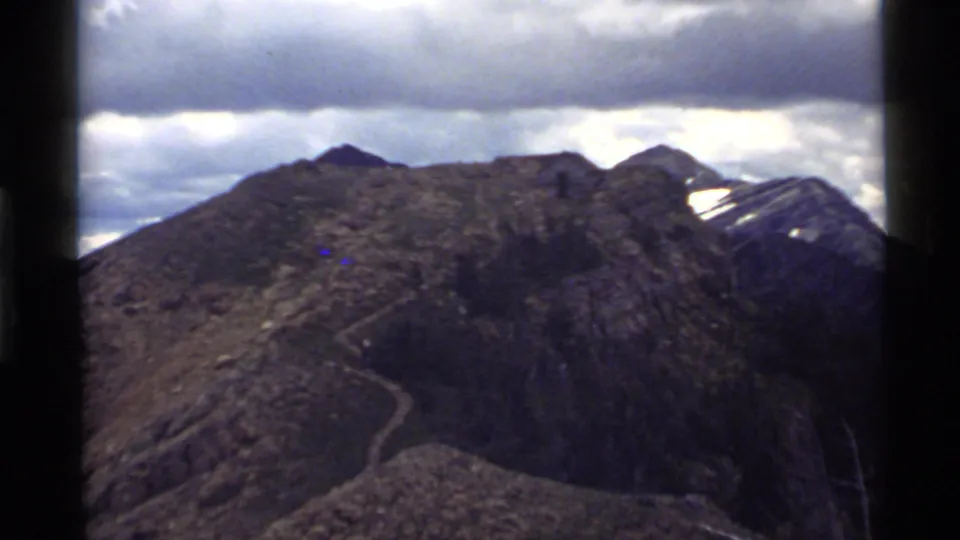 1972 8mm view of a rocky ridge near Holland Lake, Montana, with a faint trail and cloudy sky framed by film borders.