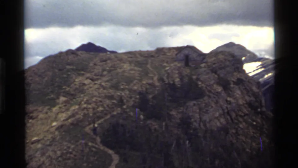 Vintage 8mm frame of a rocky Holland Lake mountain slope, with a winding trail and pale overcast sky above.