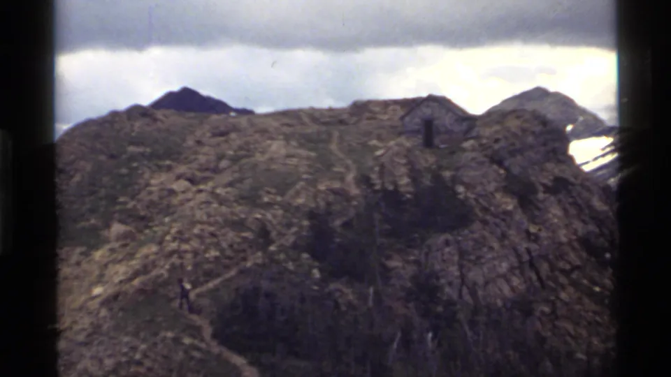 1972 8mm Holland Lake mountain view with a distant hiker on the trail, rocky ridge, snow patches, and overcast sky.