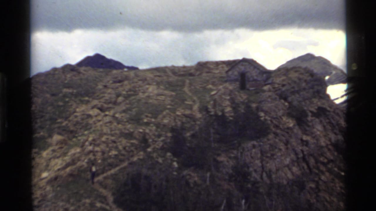 1972 Holland Lake, Montana home-movie frame showing a rocky ridge, small stone shelter, distant peaks, and overcast sky.