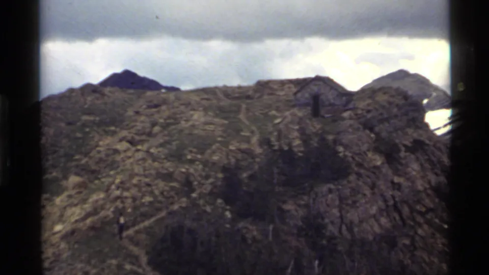 1972 Holland Lake, Montana home-movie frame showing a rocky ridge, small stone shelter, distant peaks, and overcast sky.