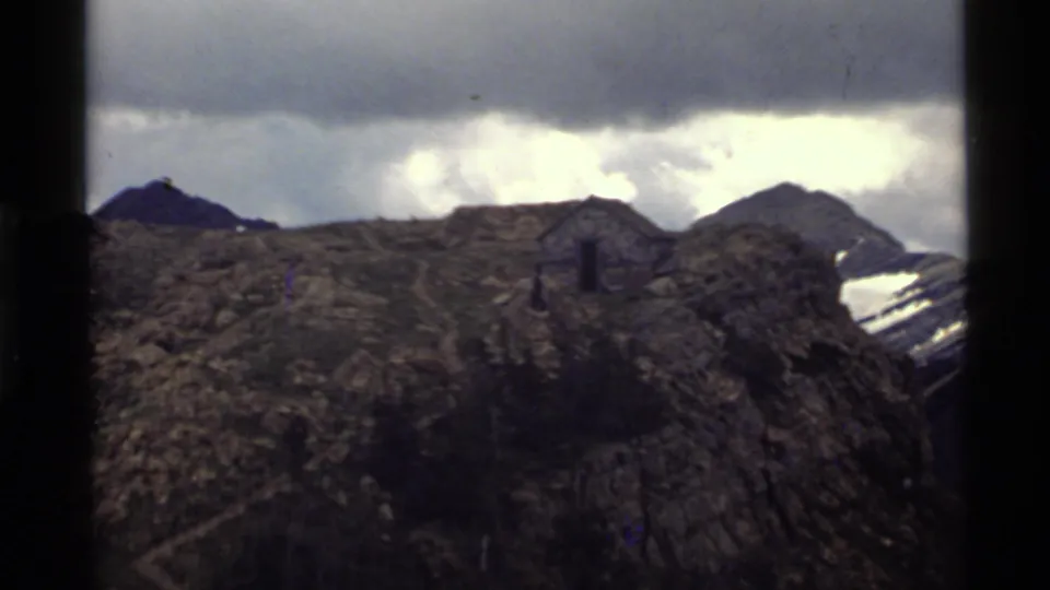The 1972 home-movie frame shows a small hiker on a ridge trail near Holland Lake, with a stone shelter and snow-streaked mountain beyond.