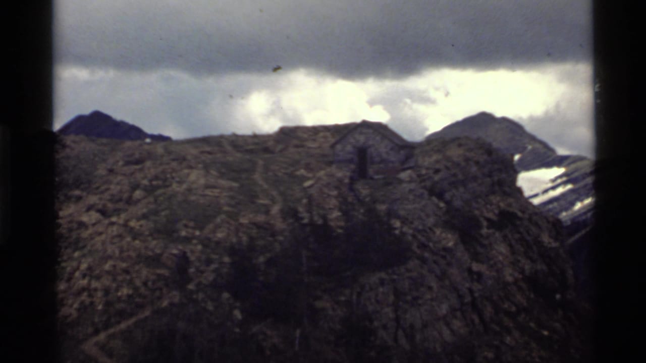 1972 Holland Lake, Montana 8mm frame showing a dark rocky ridge, small cabin-like structure, snow patches, and overcast sky.