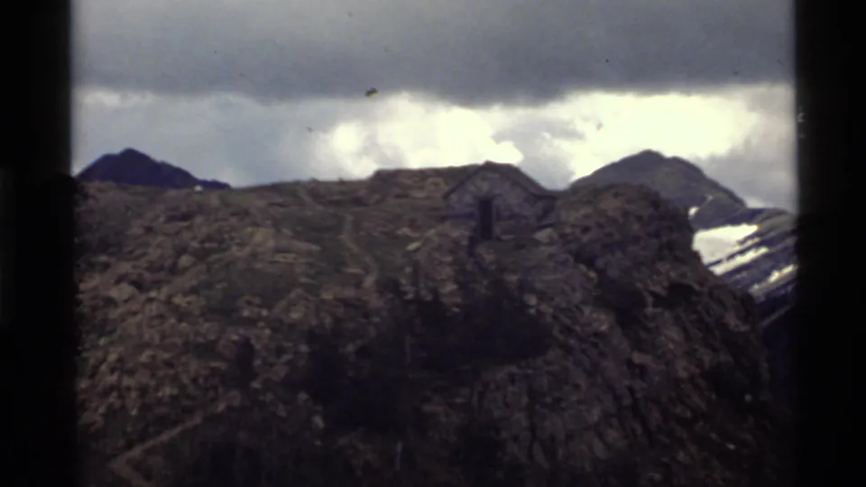 1972 Holland Lake, Montana 8mm frame showing a dark rocky ridge, small cabin-like structure, snow patches, and overcast sky.