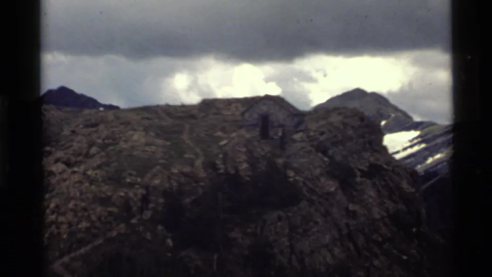 1972 Holland Lake, Montana 8mm view of a rocky overlook, small structure, dark slopes, and bright overcast cloud break.