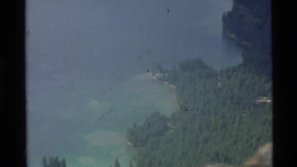 1972 Holland Lake, Montana frame showing a misty turquoise lake, evergreen forest, shoreline, and steep cliff edge from above.