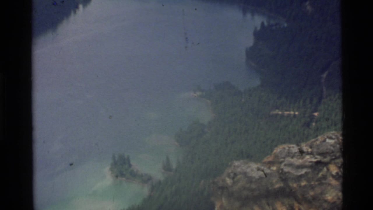 Blue-green mountain lake seen from a rocky overlook with forested shoreline and 8mm frame borders.