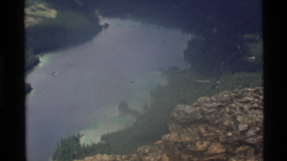 1972 Holland Lake view from above, with a rocky ledge, forested shore, coves, and soft faded 8mm texture.