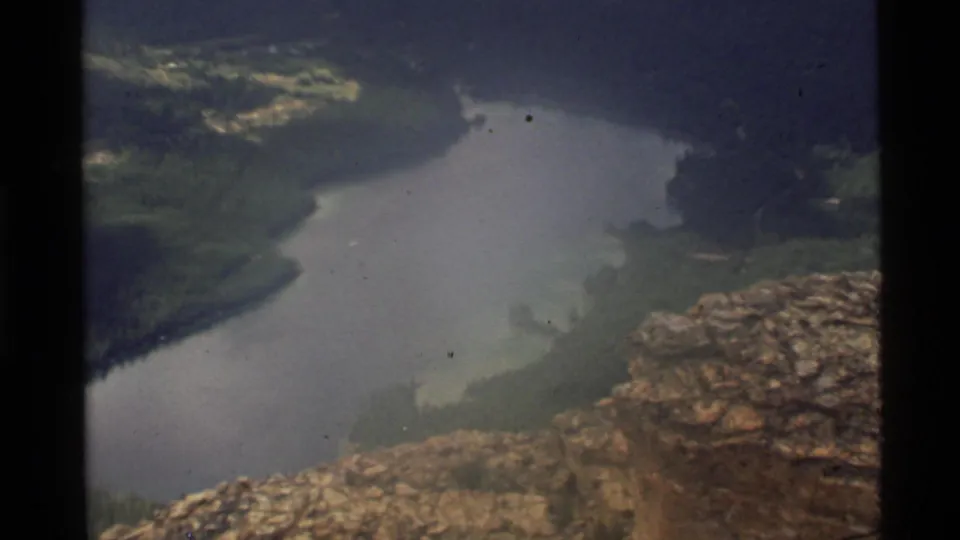 Wide 1972 home-movie view of Holland Lake, Montana, framed by rocky foreground, forested banks, and muted 8mm color.