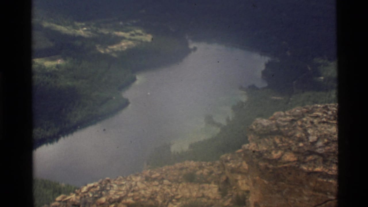 1972 8mm view over Holland Lake, Montana, seen from a high rocky overlook with dark evergreen slopes around the water.