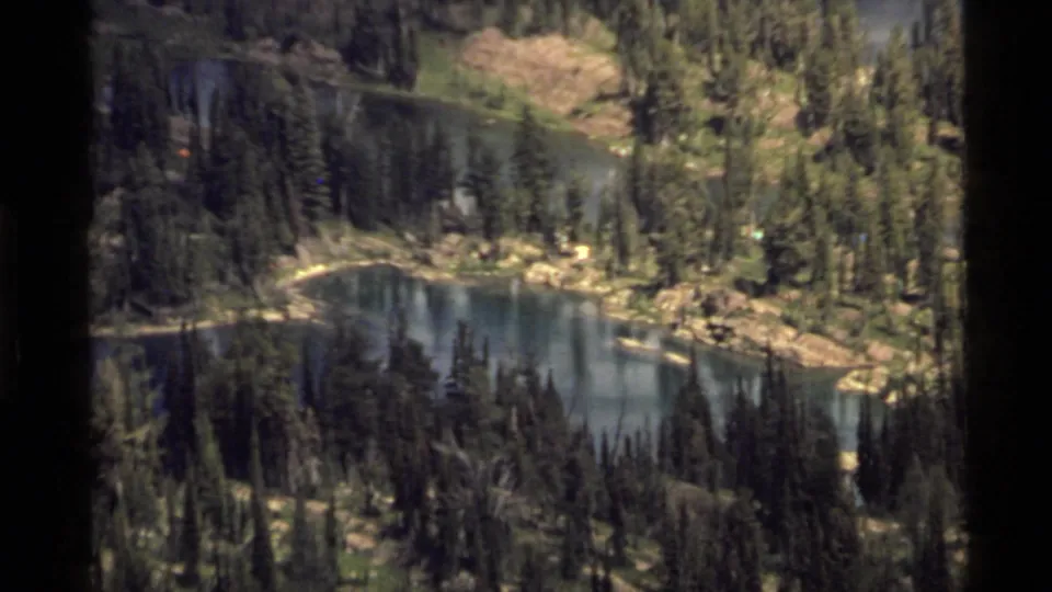 1972 8mm frame of a smaller lake basin near Holland Lake, Montana, surrounded by dense evergreens and rocky terrain.