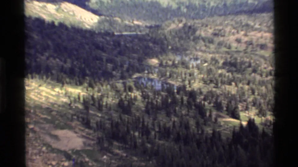 1972 home-movie aerial view over Holland Lake, Montana terrain, with evergreen forest, water, and distant rocky slopes.