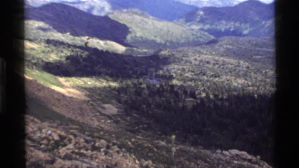 Soft 1972 8mm frame from Holland Lake, Montana, showing forested slopes, shrubland, and distant mountains from above.